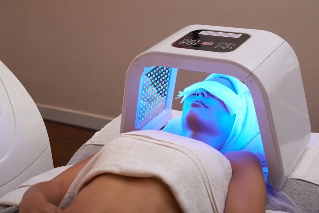 A patient at Total Skin relaxes on a treatment bed as they undergoing their blue light therapy as an acne treatment near Moorhead. The blue light device sits above and around their face and head, emitting the special colored light to help reduce inflammation.
