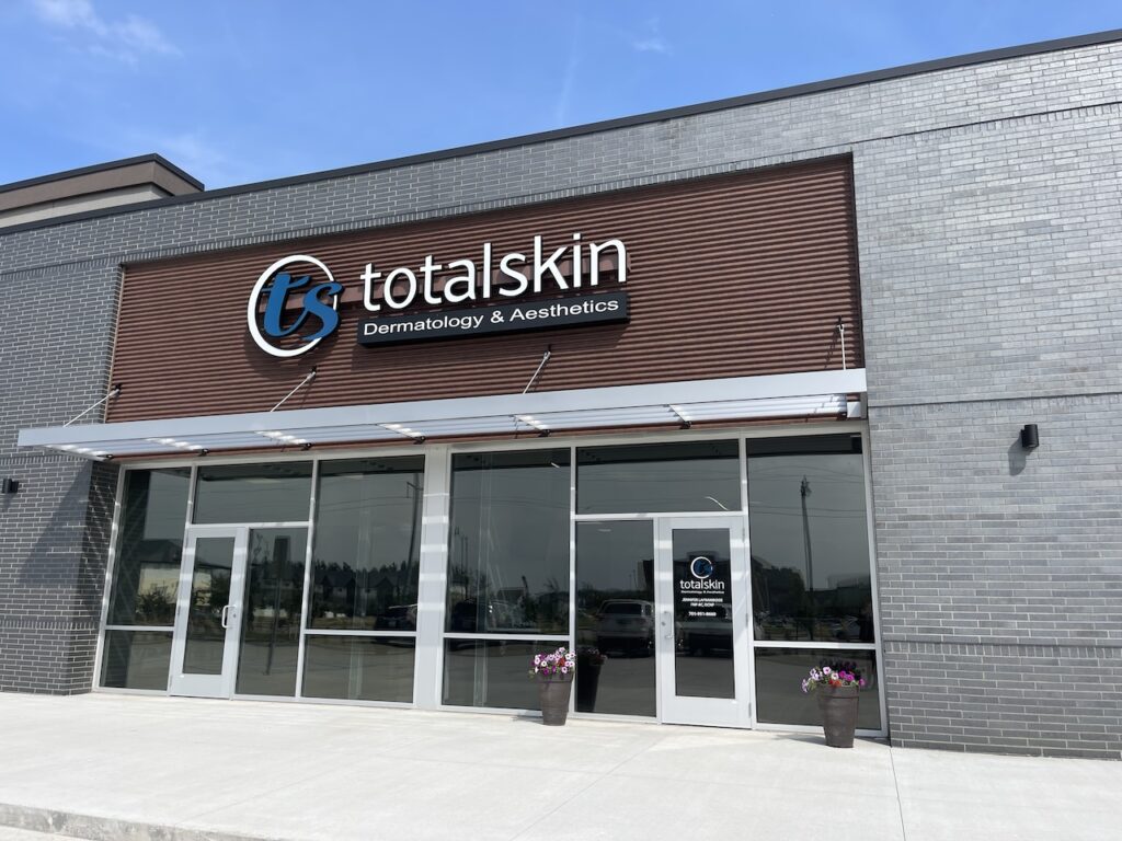 The front entrance of Total Skin, a proud weight loss clinic in Fargo. The front door sits between large, glass windows, with landscaping plants on each side. The Total Skin logo sits above the door.